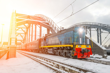 Diesel shunting locomotive overtakes passenger cars through the railway bridge in winter snow.