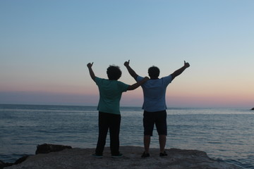 Fototapeta premium two young men thumb up near sea, background is sunset