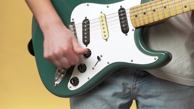 Cropped photo of young male musician in jeans tuning electric guitar before playing, isolated over yellow background