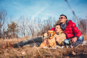 Fototapeta premium Gay male couple laying on blanket in grass with dog