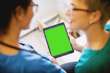 Rear view of professional middle aged nurse holding a tablet with blank editable green screen in a doctors office with woman patient in front.
