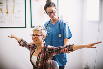 Focused professional middle aged nurse holding and checking woman patient elbows while she sitting on a chair in front with open arms.