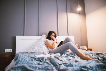 Tired sick middle aged woman holding paper wipes and a tablet while lying on the bed.