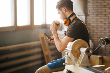 Carpenter Man using smart phone and holding cup of coffee in front of table with tools at workshop