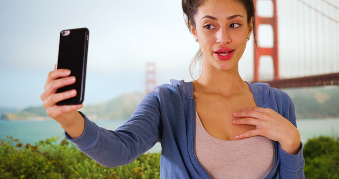 A Hispanic woman takes a selfie near the Golden Gate Bridge. A Latina millennial girl takes a picture of herself in San Francisco