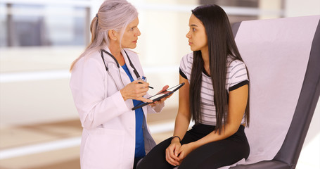 An elderly medical professional talks to her patient while using a tablet. An older doctor gives a medical examination while using her pad. 