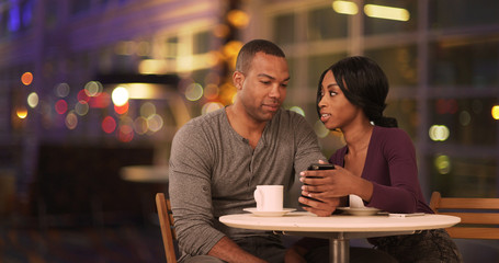 Happy Black couple using smart phone in coffee shop at night. African American man and woman using cellphone while drinking in cafe during evening. Millennials dating