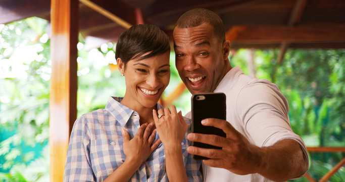 A black couple takes a selfie to celebrate their engagement. An African American man and woman take a picture using a smart phone of her new wedding ring