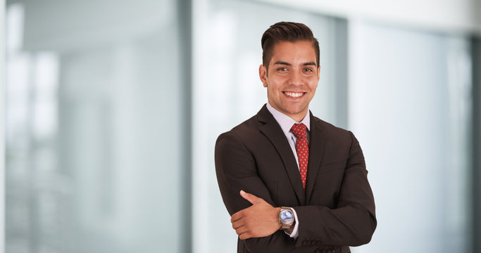 Happy smiling young Hispanic businessman standing in office with arms crossed looking at camera. Copyspace or copy space next to business professional with smile on his face