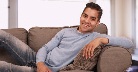 Portrait of handsome young Hispanic man sitting on couch in living room smiling at camera. Latino millennial on sofa wearing jeans and a sweater