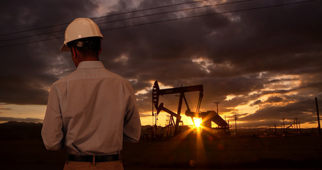 Engineer wearing hard hat helmet checking oil derrick field. Sunset as man looks at oil pump rig