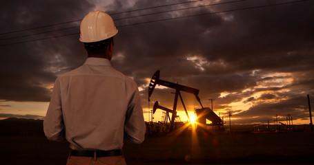 Engineer wearing hard hat helmet checking oil derrick field. Sunset as man looks at oil pump rig