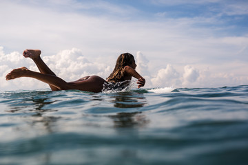 Beautiful young indonesian woman in bikini surfing wave in Bali on the background of blue sky, clouds and tropical beach - obrazy, fototapety, plakaty