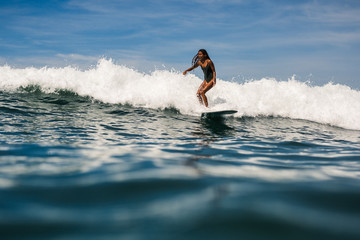 Beautiful young indonesian woman in bikini surfing wave in Bali on the background of blue sky,...