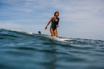 Beautiful young indonesian woman in bikini surfing wave in Bali on the background of blue sky, clouds and tropical beach