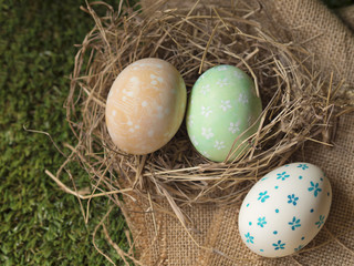 Top view of Easter eggs, yellow marble, green flower, and white with blue color flower pattern handmade painting style on bird nest and brown sack on green grass in Spring garden