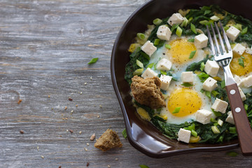 Green shakshuka with spinach, leek and feta in a ceramic frying pan on a wooden table with bread, free space, selective focus. Delicious healthy home food
