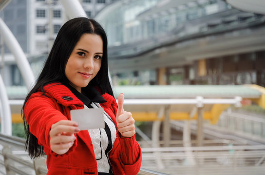 beautiful cheerful young woman in red jacket holding credit card and showing thumbs up in modern big city, fashion, promotion, special offer, winter sale, people, payment, shopping online concept