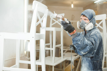 Man painting chair into white paint in respiratory mask. Application of flame retardant ensuring fire protection, airless spraying. - obrazy, fototapety, plakaty
