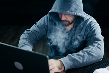 a criminal man hides his face under a hood, hacks a laptop, lots of smoke