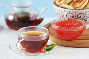 transparent glass mug with black tea and mint on a background of pancakes and caviar