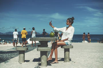 Young cute Brazilian female in white chemise is taking selfie using her cell phone while sitting on...