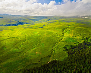 Aerial view of green hills. Summer landscape. Green grassy meadow on a hillside on top of mountain ridge with some forest under cloudy blue sky. Adygea, Lago-naki, Krasnodar, Russia.