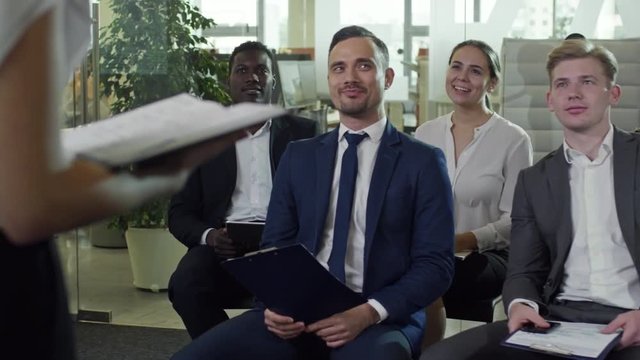 PAN of businessman in suit holding clipboard and laughing while talking to unrecognizable businesswoman delivering speech before cheerful colleagues
