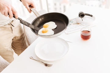 Cropped photo of man in casual clothing putting frying eggs into plate on table, while having dinner in home kitchen