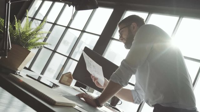 Man with a beard in white shirt works against the background of a large panoramic window, leans on table. Young businessman works alone in bright spacious office, stylish accessories and plants.