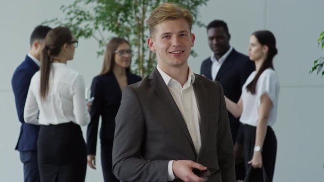 Tilt up of successful businessman in suit holding mobile phone and smiling for camera while his colleagues discussing work in background
