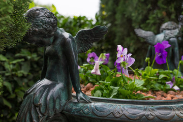 Flowerpot decorated with two little angels and purple flowers inside. Flower pot is located in a garden