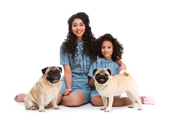 happy young mother and daughter sitting on floor with two pugs isolated on white