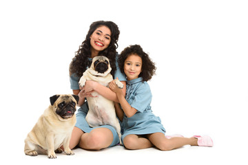 happy young mother and daughter sitting on floor with two adorable pugs isolated on white