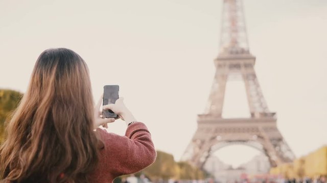 Back view of young brunette female taking photos of Eiffel tower on smartphone. Teenager exploring Paris, France.