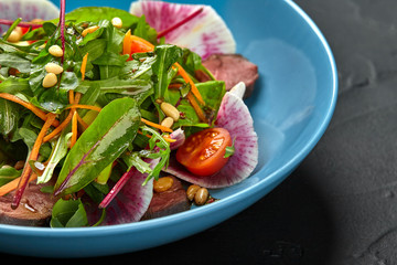 Spicy beef slices meat salad with tomatoes, parsley, radish and salad leaves spinach in a blue plate on a black table. Close-up