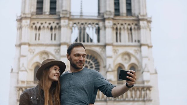 Young happy couple hugging and taking selfie photos on smartphone near the famous Notre Dame in Paris, France.