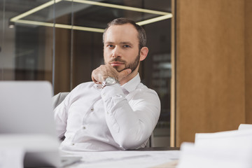 Naklejka premium handsome thoughtful architect sitting at workplace and looking at camera