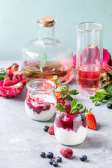 Jars of natural white yogurt with berry sauce, fruit salad with pink dragon fruit, berries and mint, served with bottle of lemonade on grey table. Healthy eating
