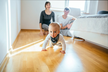 Couple sitting on the floor in bedroom with infant baby boy smiling and crawling to the camera. Happy family spending time tigehter at home in sunny day.