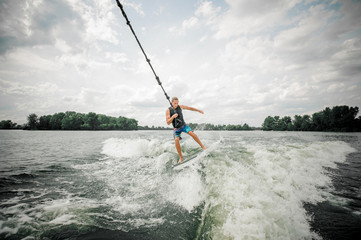 Young and athletic man wakesurfing on the board holding a cable