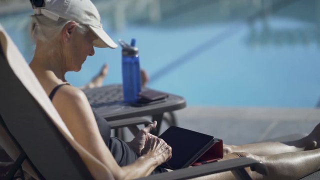 Over The Shoulder Shot Of Elderly Woman In Lounge Chair At An Outdoor Pool Reading On A Tablet Or Ipad.