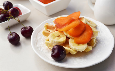 Homemade belgian waffles with fresh apricot slices, banana and sweet cherry. Delicious breakfast in a white plate.