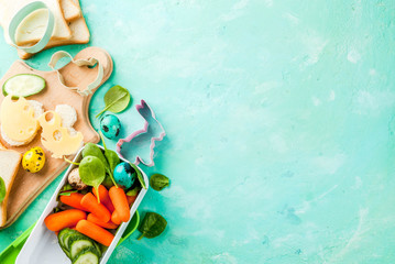 Cooking creative kids breakfast lunch box for Easter, sandwiches with cheese, fresh vegetables - cucumbers, carrots, spinach, colorful quail eggs. Light blue table, copy space top view