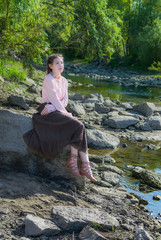 Beautiful girl dressed in Boho style, sits on a rock by the river