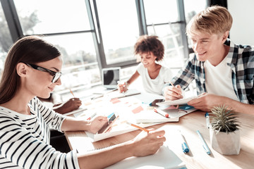 Together with colleagues. Nice positive young woman sitting at the table and writing while working together with her colleagues