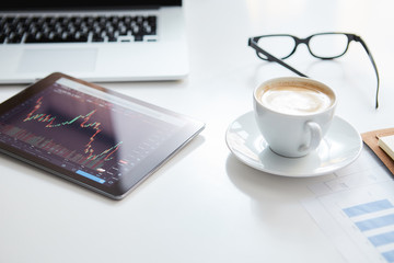 Business workplace with stock market data application on a tablet, banking interface on a tablet and some papers with charts, graphs and numbers on a desktop.