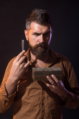 Attractive yound barber with thick black beard and mustache smiling while holding straight razor near his beard. Brutal caucasian hipster, bearded man holds in hands dangerous razor, wooden casket.
