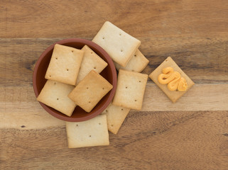 Top view of a serving of hard bread crackers in a red clay bowl with one to the side having canned cheese on a wood table.
