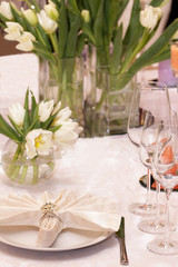 A stylish modern wedding decor of dishes, white bouquets with flowers, candles and glasses stand on a light tablecloth of a served table in a restaurant. Side view, selective focus